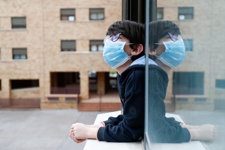 Un neno cunha máscara asómase á xanela da súa casa durante o confinamento polo coronavirus 